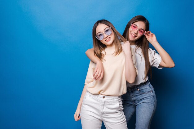 Amigos para siempre. Dos amigas encantadoras lindas en gafas de sol posando con una sonrisa aislada en la pared azul