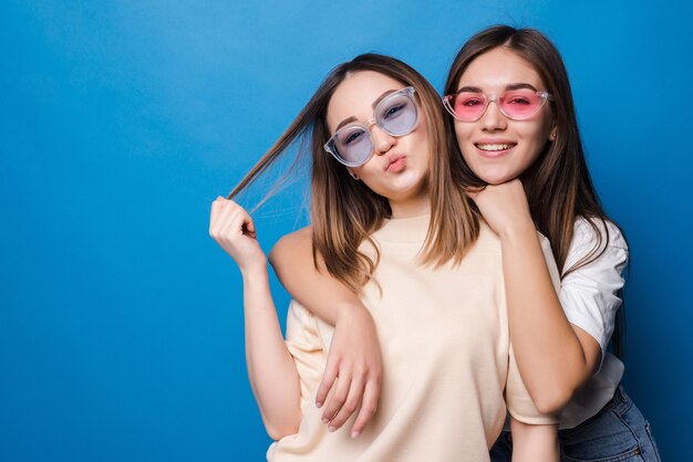 Amigos para siempre. Dos amigas encantadoras lindas en gafas de sol posando con una sonrisa aislada en la pared azul