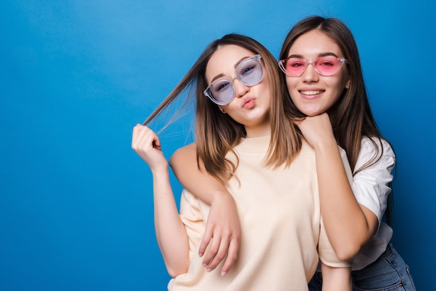 Amigos para siempre. Dos amigas encantadoras lindas en gafas de sol posando con una sonrisa aislada en la pared azul