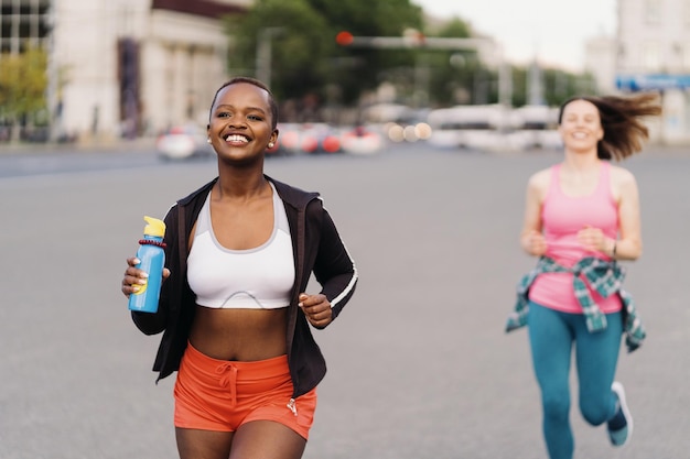 Amigos sonrientes alegres en ropa deportiva corriendo en la ciudad hablando de mujeres multiétnicas haciendo ejercicio físico