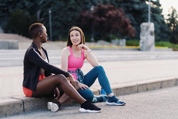 Amigos sonrientes alegres en ropa deportiva sentados en la ciudad hablando de mujeres multiétnicas que tienen un descanso de entrenamiento físico