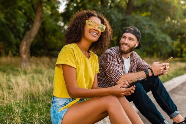 Amigos sonrientes jovenes felices que se sientan parque usando smartphones, hombre y mujer que se divierten juntos