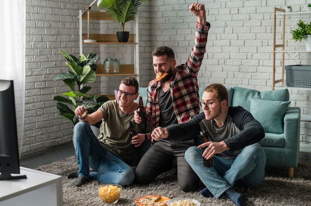 Amigos varones alegres viendo deportes en la televisión y comiendo pizza