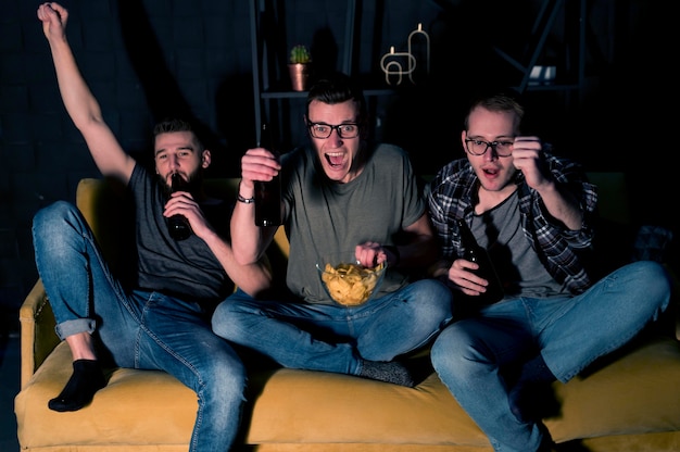 Amigos varones alegres viendo deportes en la televisión juntos mientras toman bocadillos y cerveza