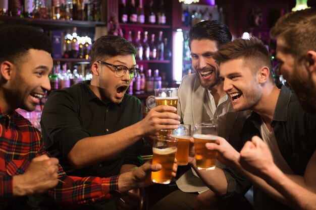 Amigos varones felices tintinean con jarras de cerveza en pub