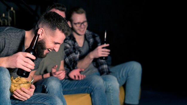 Amigos varones sonrientes viendo deportes en la televisión juntos mientras toman bocadillos y cerveza