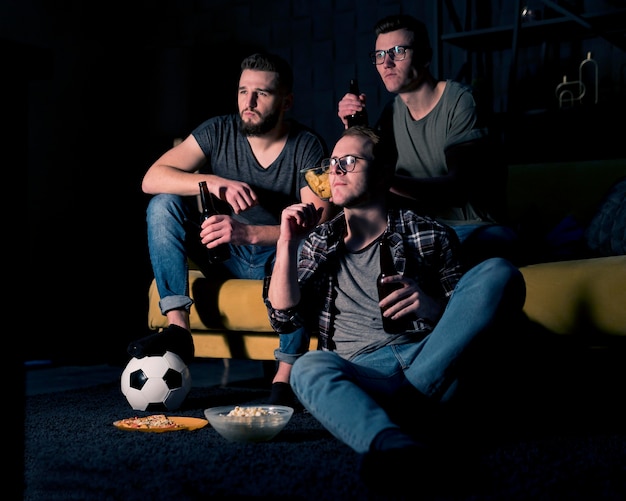 Amigos varones viendo deportes en la televisión juntos mientras toman cerveza y bocadillos