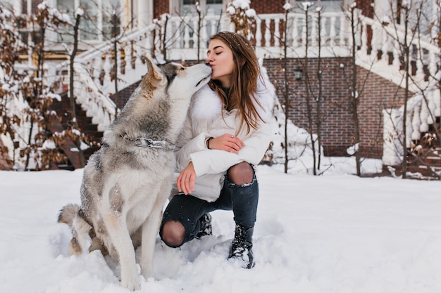 Amistad real, encantadores momentos felices de una encantadora joven con un lindo perro husly disfrutando del frío invierno en la calle llena de nieve. Mejores amigos, amor de los animales, emociones verdaderas, dar un beso.