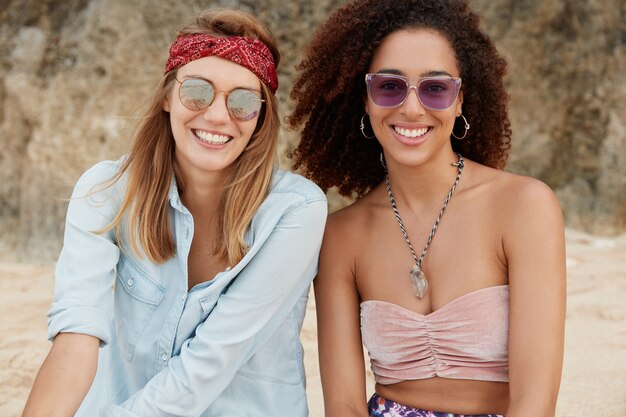 Las amistosas mujeres interraciales usan gafas de sol de moda y ropa de verano, están de buen humor al respirar el aire fresco del mar, tienen expresiones felices, se sientan en la playa de arena.