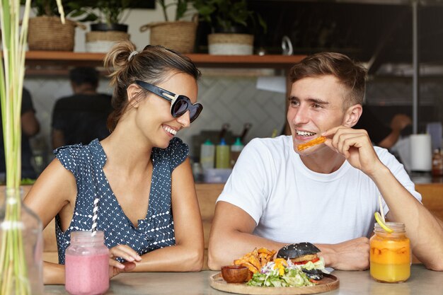 Amor y amistad. Pareja feliz comiendo hamburguesas con papas fritas y tomar bebidas frescas durante la fecha en la acogedora cafetería. Linda mujer en gafas de sol de moda escuchando los chistes de su novio y riendo