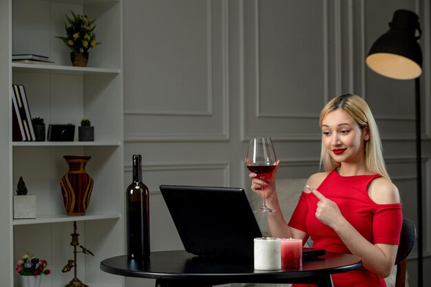 Amor virtual linda chica rubia con vestido rojo en una cita a distancia con vino apuntando a la copa de vino