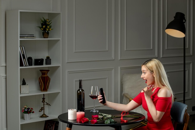 Amor virtual linda chica rubia con vestido rojo en cita a distancia con vino emocionado en cámara