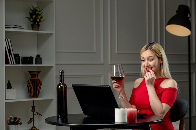 Amor virtual linda chica rubia en vestido rojo en cita a distancia con vino mordiendo el dedo