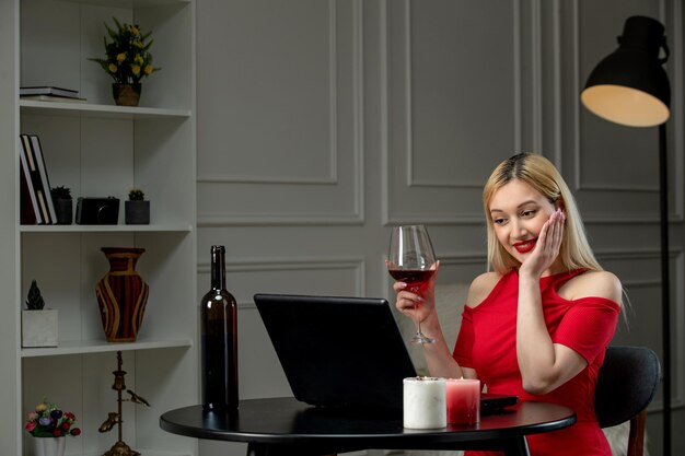 Amor virtual linda chica rubia con vestido rojo en una cita a distancia con vino tomados de la mano en la cara