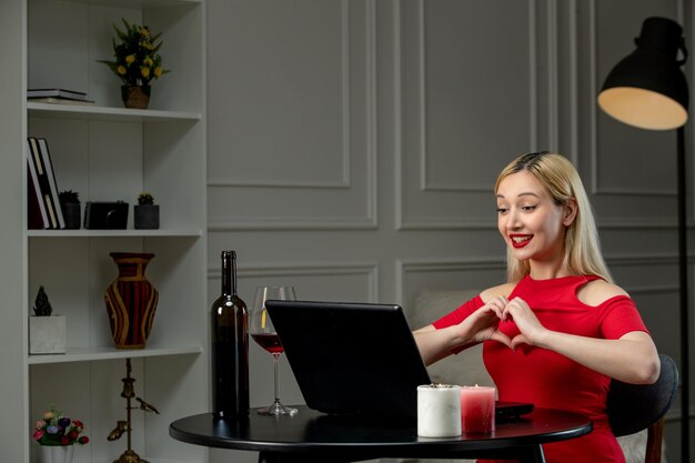 Amor virtual linda chica con vestido rojo en una cita a distancia con vino que muestra el signo del corazón