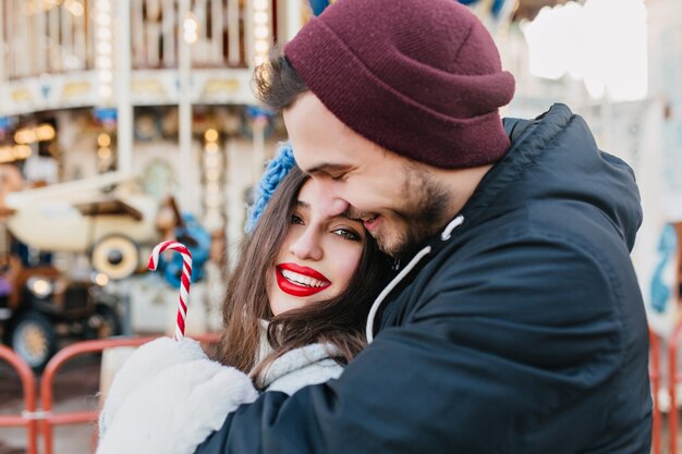 Amorosa pareja abrazándose en el parque de atracciones en fin de semana de invierno. Feliz niña de pelo negro celebrando la Navidad con su novio y posando delante del carrusel en un día frío.