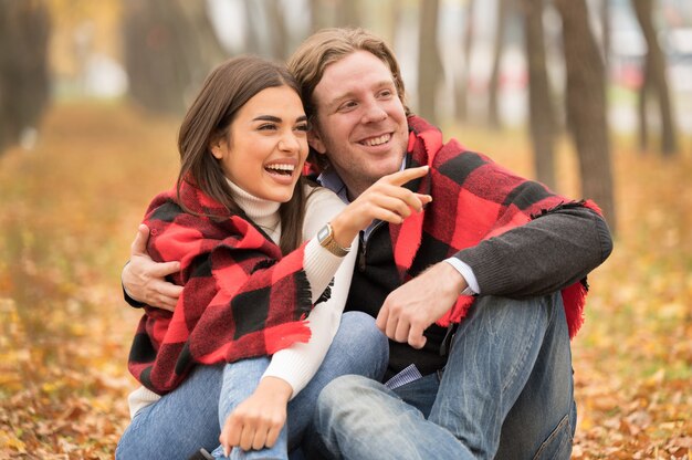 Amorosa pareja caucásica abrazándose con una manta sobre ellos sentado en el bosque de otoño