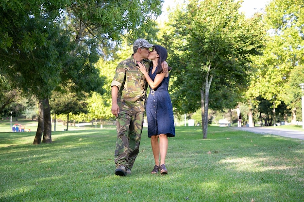 Amorosa pareja caucásica abrazar, besar y caminar juntos sobre el césped en el parque. Soldado de mediana edad con uniforme militar, abrazando a su bella esposa. Concepto de reunión familiar, fin de semana y regreso a casa