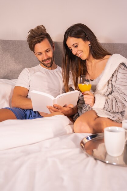 Amorosa pareja caucásica bebiendo jugo y café en la cama mientras lee un libro en una habitación de hotel