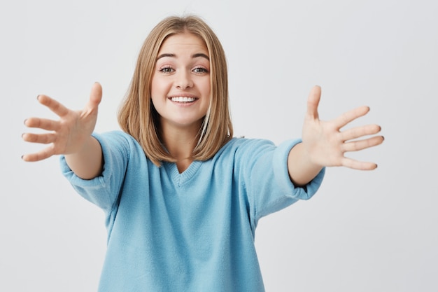 Ampliamente sonriendo con dientes bastante joven mujer europea vestida con suéter azul extendiendo sus brazos, posando. Emociones y sentimientos positivos.