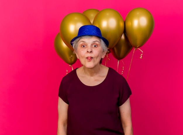 Anciana alegre con gorro de fiesta se encuentra frente a globos de helio atasca la lengua mirando a la cámara en rosa