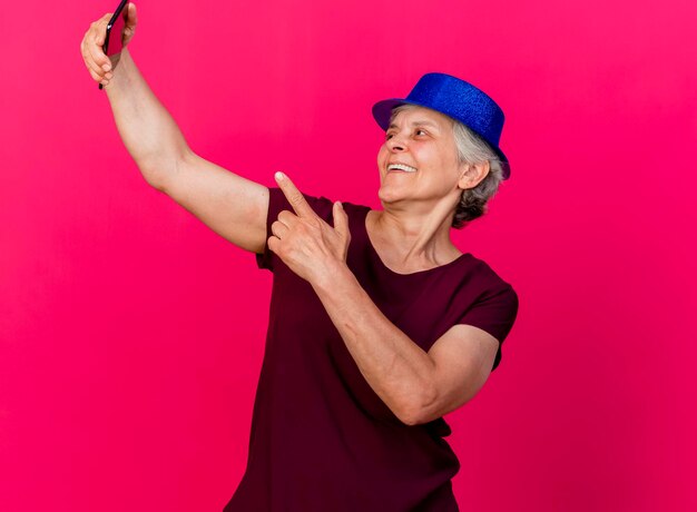 Anciana alegre con gorro de fiesta mira y apunta al teléfono en rosa