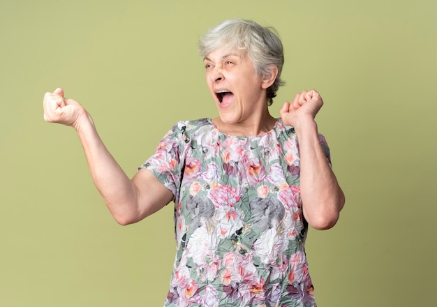 Anciana alegre levanta los puños mirando al lado aislado en la pared verde oliva