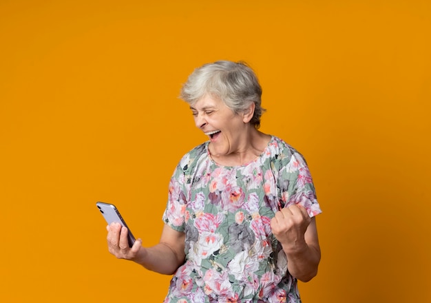 Foto gratuita anciana alegre mira teléfono sosteniendo puño aislado en pared naranja