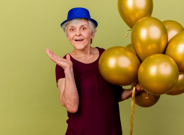 Anciana alegre con sombrero de fiesta se encuentra con globos de helio sosteniendo la mano abierta en verde oliva