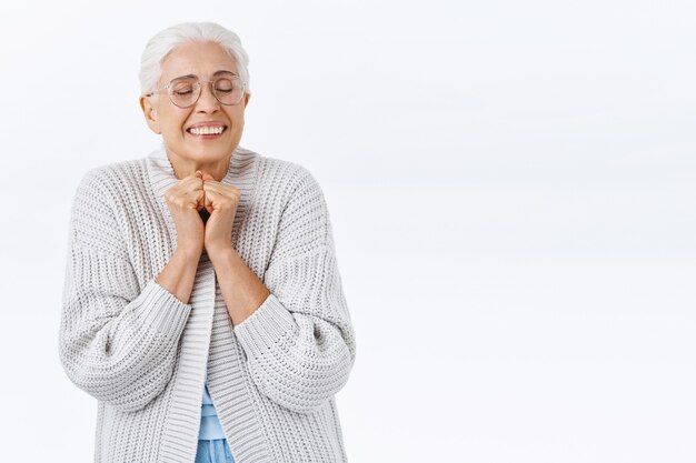 Una anciana alegre y soñadora que siente afecto y alegría al recordar un bonito recuerdo, juntar las manos cerca del pecho, cerrar los ojos y sonreír a los niños emocionados y complacidos que regresan a casa para las vacaciones.