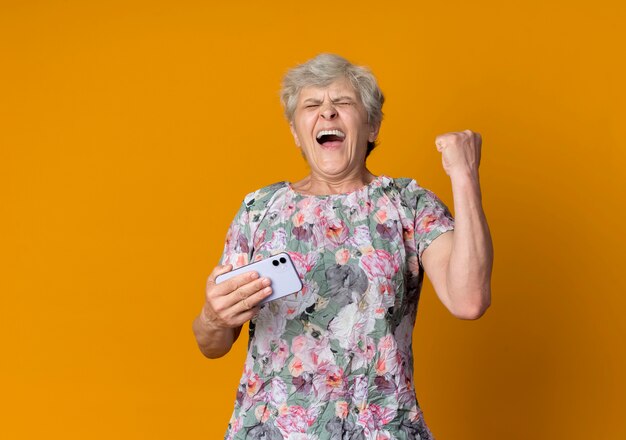 Anciana alegre sostiene el teléfono y levanta el puño aislado en la pared naranja