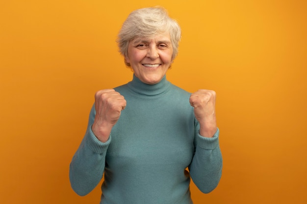 Anciana alegre vistiendo suéter de cuello alto azul apretando los puños haciendo un gesto fuerte