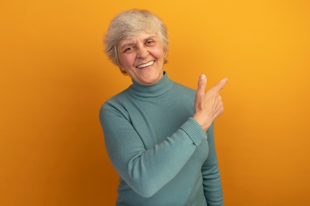 Foto gratuita anciana alegre vistiendo suéter de cuello alto azul apuntando al lado aislado en la pared naranja con espacio de copia