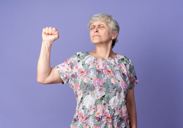 Anciana ansiosa levanta el puño con los ojos cerrados aislado en la pared púrpura