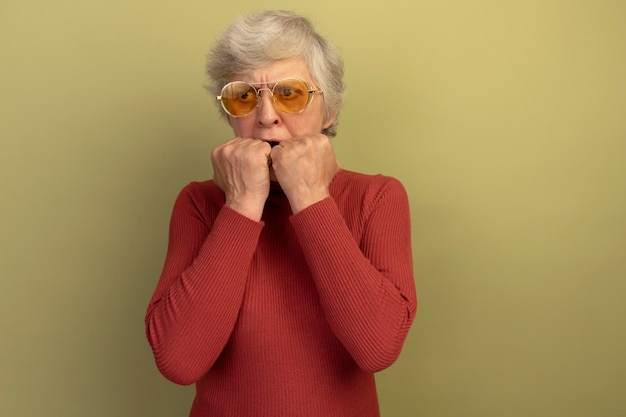 Anciana ansiosa con suéter de cuello alto rojo y gafas de sol mirando al lado manteniendo las manos en la boca aisladas en la pared verde oliva con espacio de copia