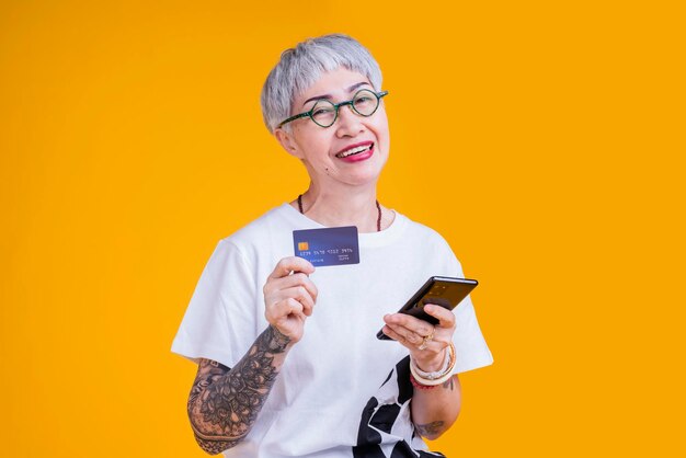 Anciana asiática mayor tatuaje mano mostrar tarjeta de crédito y teléfono inteligente con sonrisa confiada y alegre asia anciana cabello gris de pie gesto de la mano mantener atención de crédito pago sin efectivo ideas de negocios