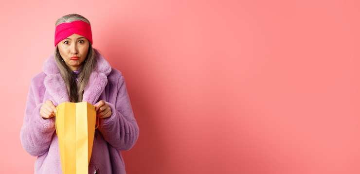 Foto gratuito una anciana asiática molesta abre una bolsa de compras y mira triste a la cámara de pie sobre un fondo rosa