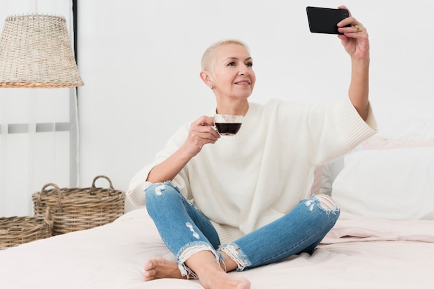 Anciana en la cama sosteniendo la taza de café y tomando selfie