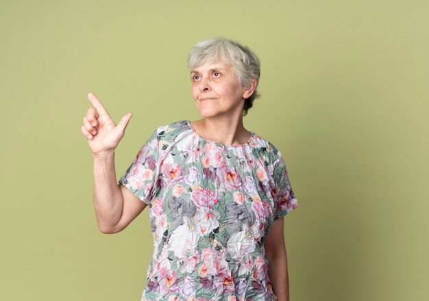 Foto gratuita anciana complacida apunta al lado mirando hacia arriba aislado en la pared verde oliva