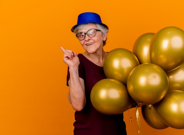 Anciana complacida en gafas ópticas con sombrero de fiesta se encuentra con globos de helio apuntando al lado aislado en la pared naranja