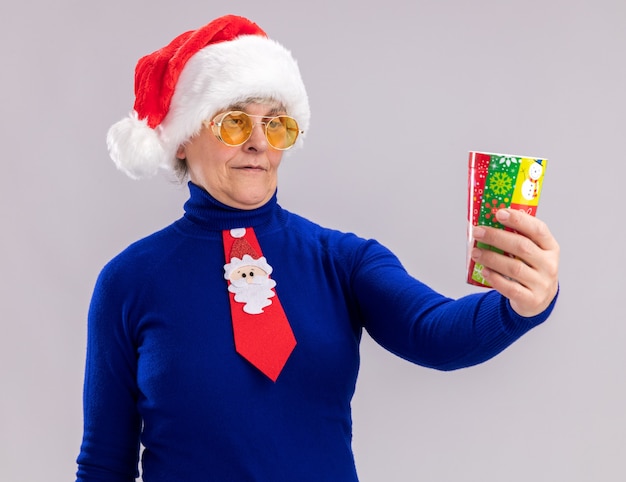 Anciana complacida en gafas de sol con gorro de Papá Noel y corbata de santa sosteniendo y mirando el vaso de papel aislado en la pared blanca con espacio de copia