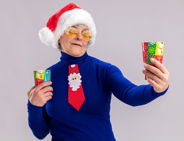 Anciana complacida en gafas de sol con gorro de Papá Noel y corbata de santa sosteniendo y mirando vasos de papel aislados en la pared blanca con espacio de copia