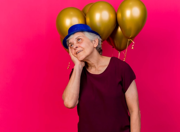 Anciana complacida con gorro de fiesta se encuentra frente a globos de helio poniendo la mano en la cara en rosa