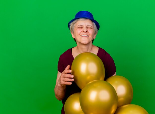 Anciana complacida con gorro de fiesta se encuentra con globos de helio aislados en la pared verde con espacio de copia