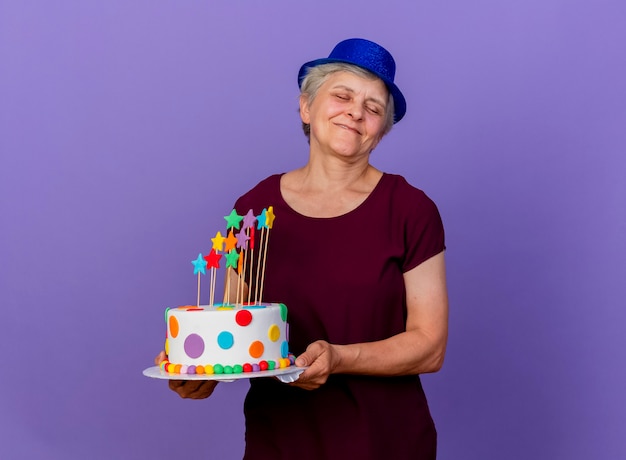 Anciana complacida con gorro de fiesta tiene pastel de cumpleaños aislado en la pared púrpura