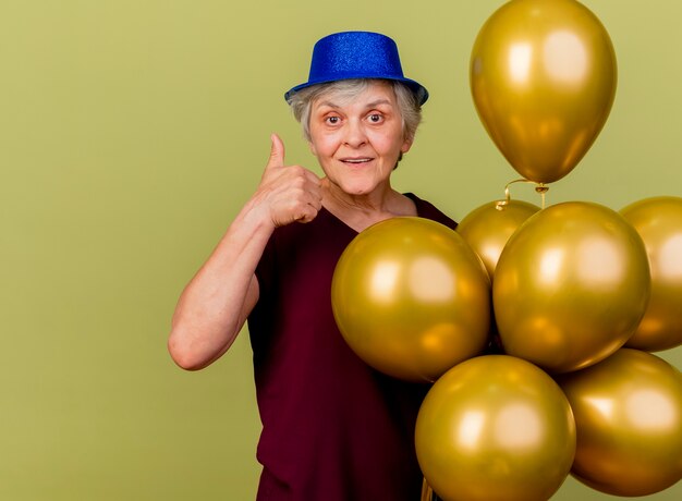 Anciana complacida con sombrero de fiesta se encuentra con globos de helio y pulgares arriba aislados en la pared verde oliva con espacio de copia