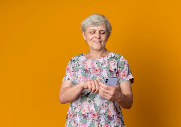 Anciana complacida sosteniendo y mirando el teléfono aislado en la pared naranja
