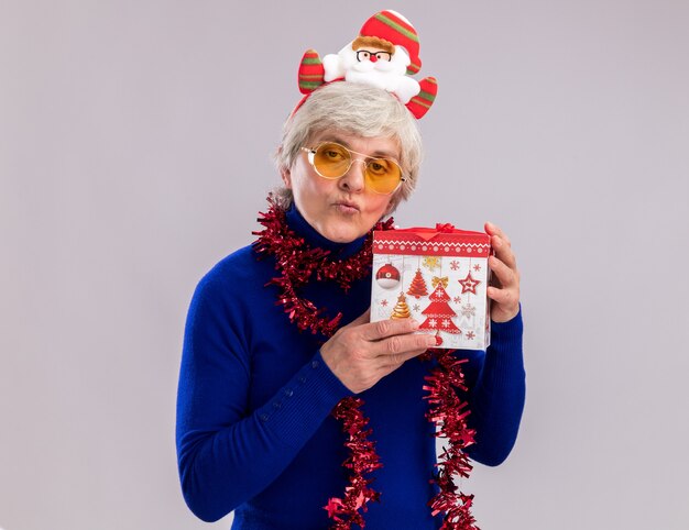 Anciana confiada en gafas de sol con diadema de santa y guirnalda alrededor del cuello tiene caja de regalo de Navidad aislada en la pared blanca con espacio de copia