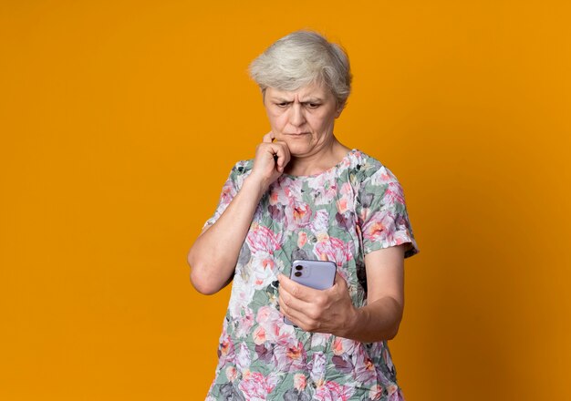 Anciana confundida pone la mano en la barbilla mirando el teléfono aislado en la pared naranja