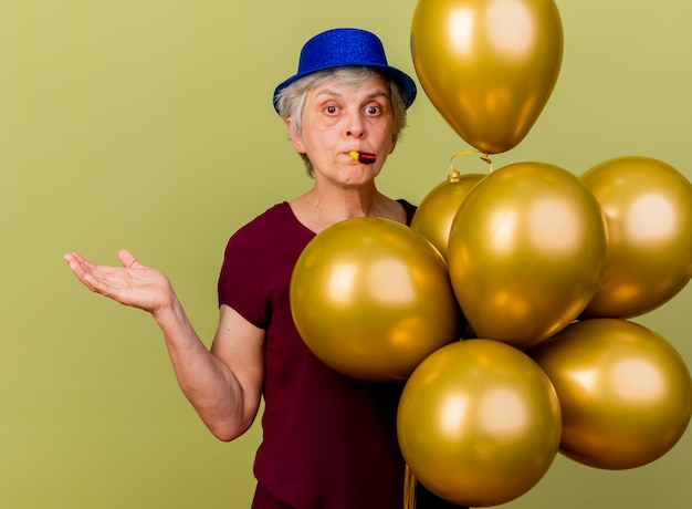 Anciana confundida con sombrero de fiesta se encuentra con globos de helio sostiene la mano abierta aislada en la pared verde oliva con espacio de copia